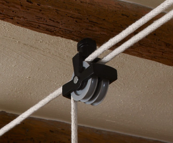 A black double screw pulley installed into a wooden beam. The sash cord runs through the pulley and out the other side. 